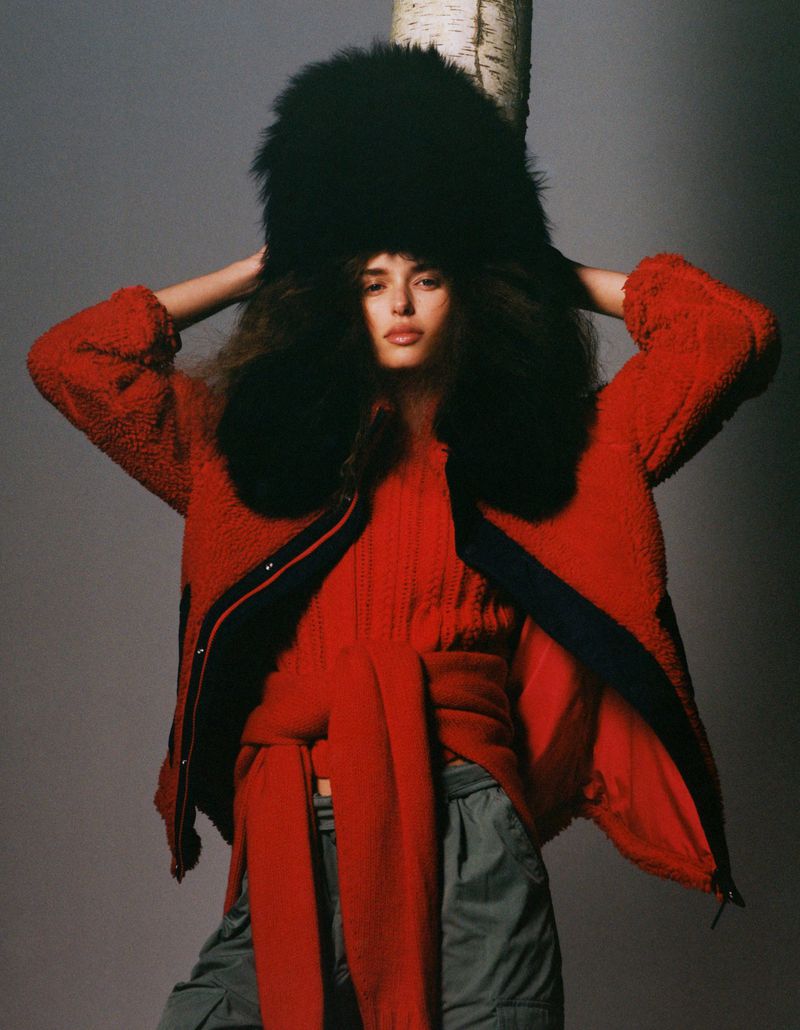 Fashion shot of a model in a red jacket, matching sweater, and fur hat, standing in front of a birch tree with a grey background.