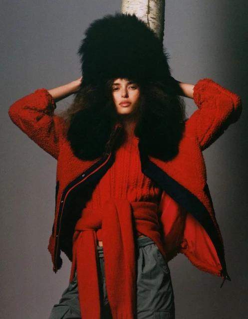 Fashion shot of a model in a red jacket, matching sweater, and fur hat, standing in front of a birch tree with a grey background.