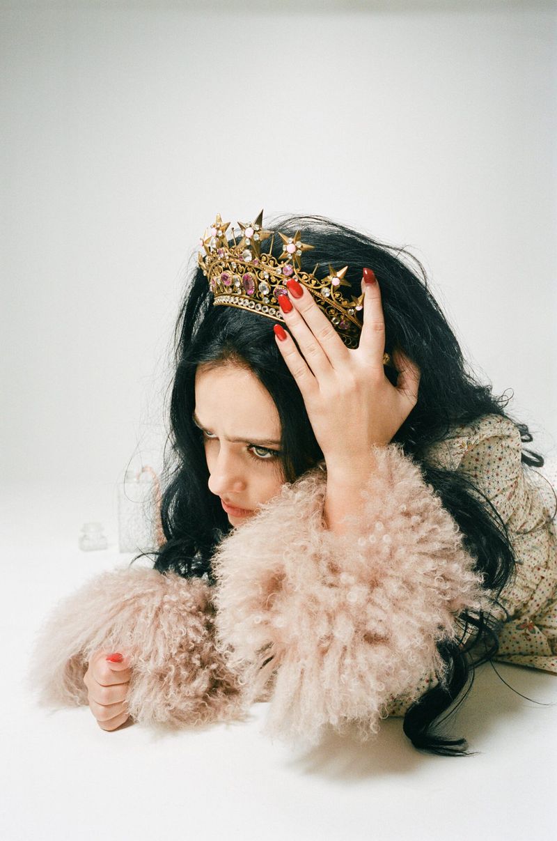 Portrait of a woman with dark hair and red nails wearing a jeweled gold crown and a shaggy pink jacket against a white background.