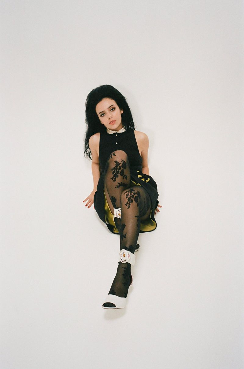 Young woman in a black dress, black patterned stockings, and white heels posing against a white background in a studio setup.