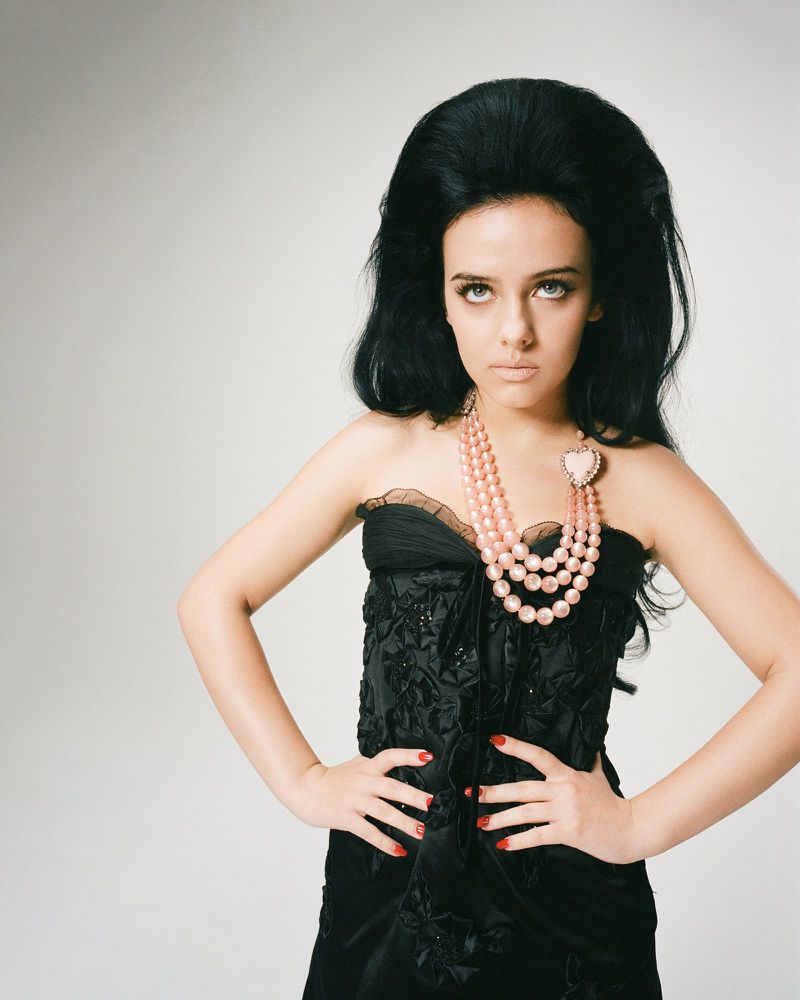 A woman with dark hair and a pink beaded necklace poses indoors. She wears a black strapless dress with a decorative floral pattern.