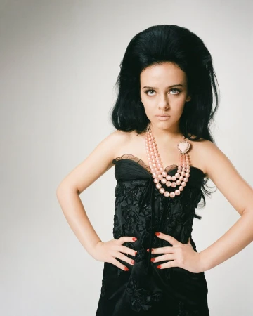 A woman with dark hair and a pink beaded necklace poses indoors. She wears a black strapless dress with a decorative floral pattern.