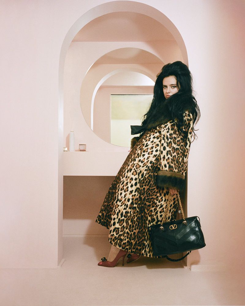 A woman in a leopard print coat poses in a pink interior with archways, holding a black Valentino purse with gold chain straps.