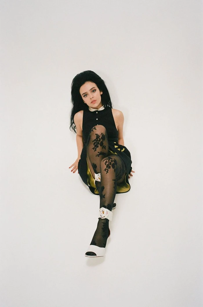Jessie Murph sitting on a white studio floor wearing a black dress and patterned tights for Interview Magazine
