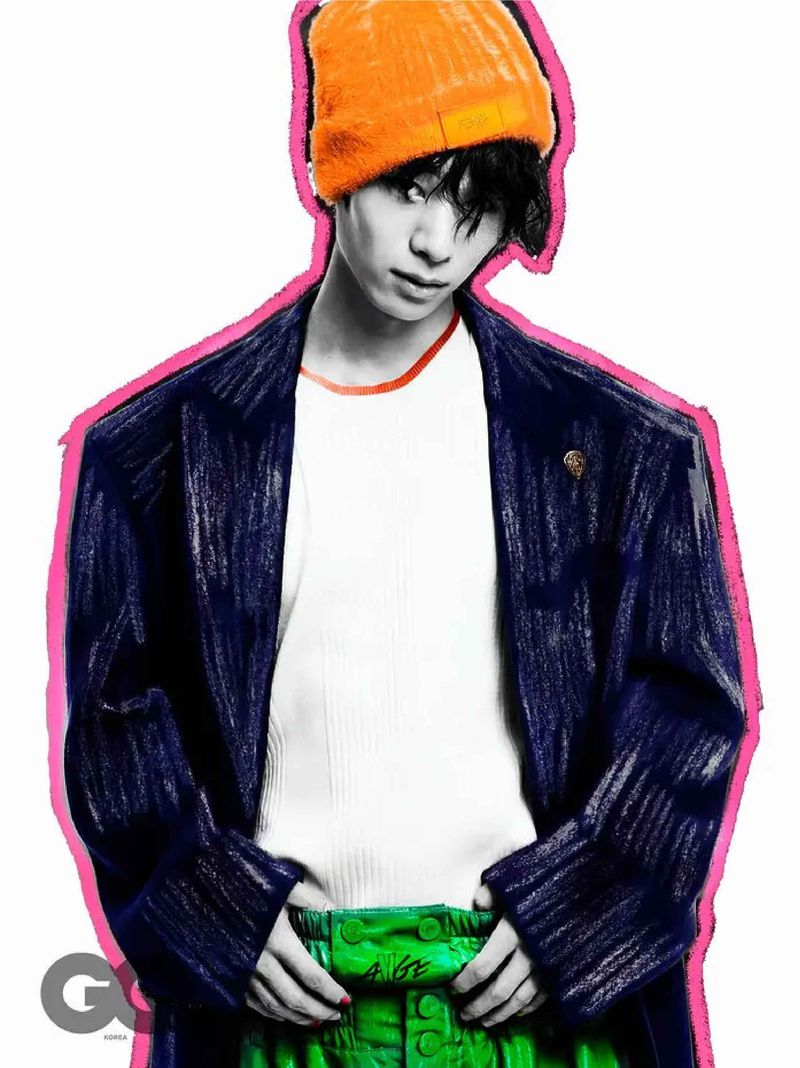 Person in an orange knit cap, a dark-blue blazer, and green pants with an artistic border, facing the viewer with a serious look.