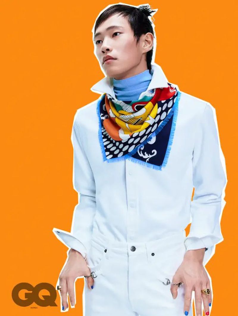 Portrait of an adult male model with dark hair posing in a white shirt, pants, and colorful scarf, standing against an orange background.