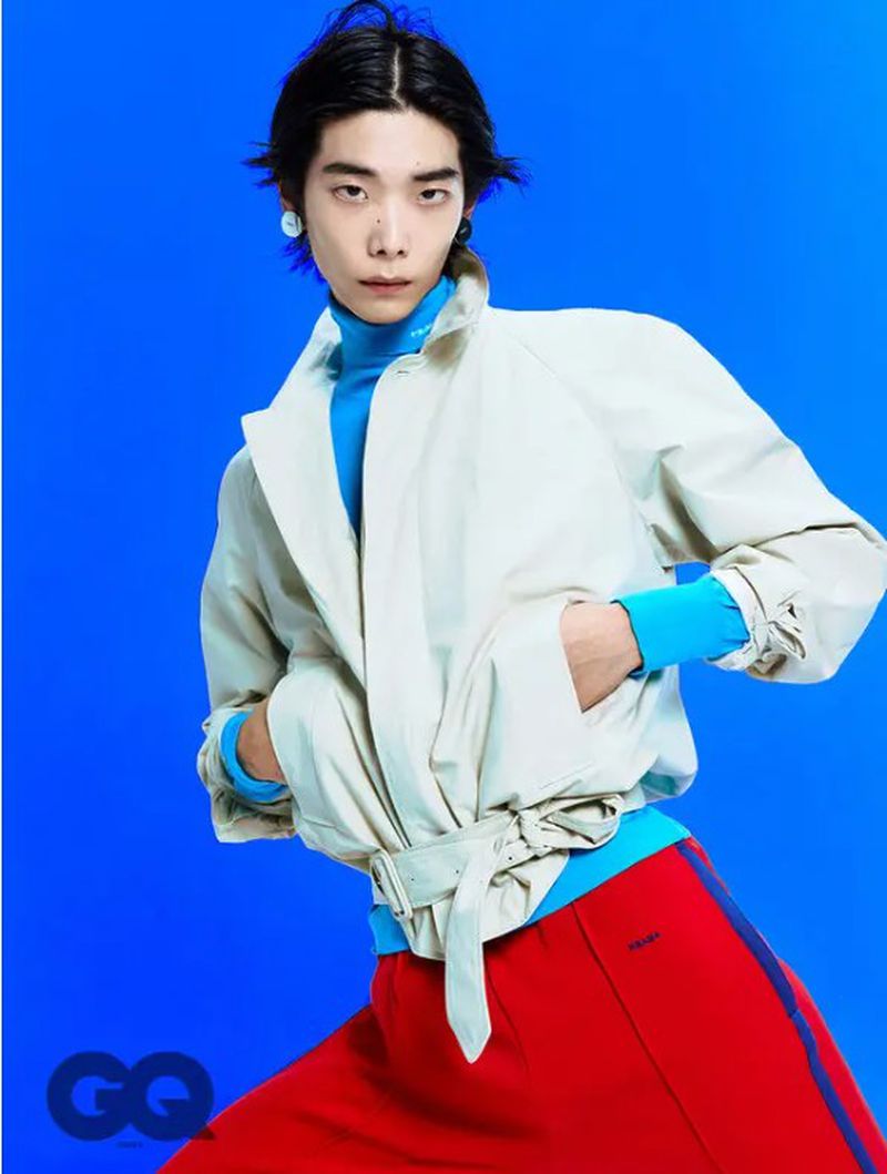 Fashion portrait of a young Asian man in a light jacket, blue turtleneck, and red pants against a vibrant blue background.