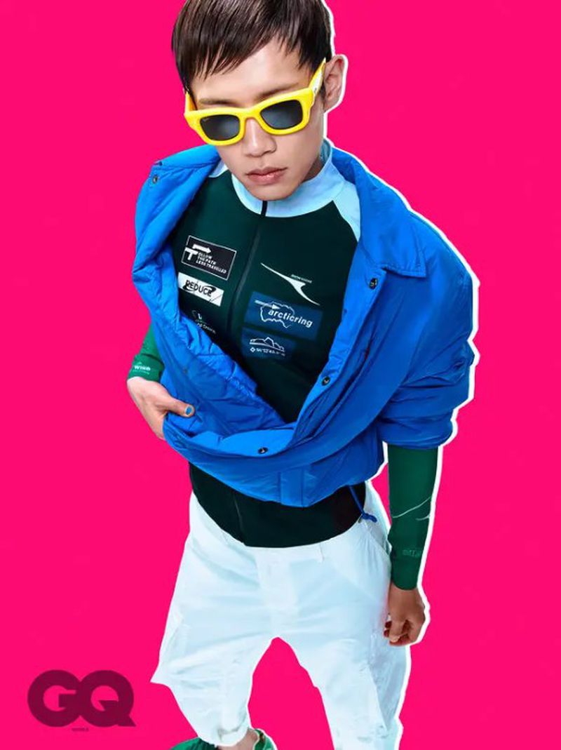 Stylish young man in blue jacket and yellow sunglasses poses against a vibrant pink backdrop in a fashion editorial shot.