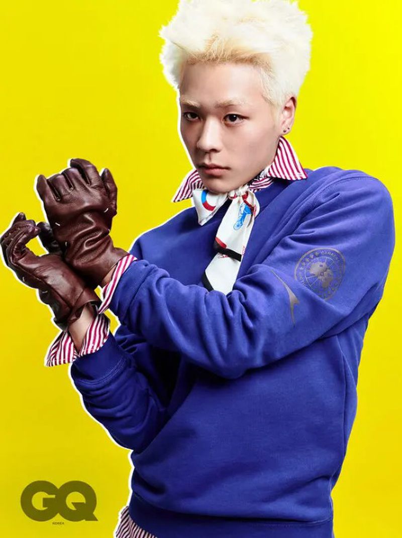 Stylish blonde male poses in a blue sweatshirt, brown gloves, and neck scarf against a bright yellow backdrop with magazine logo.