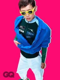 Model wearing yellow sunglasses, blue jacket, green athletic top, and white pants against a bright pink background.