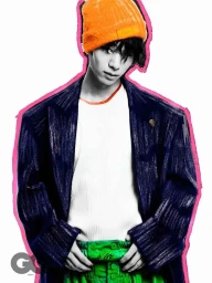Portrait of a man wearing a bright orange beanie, oversized dark coat, white t-shirt, and green trousers against a white backdrop.