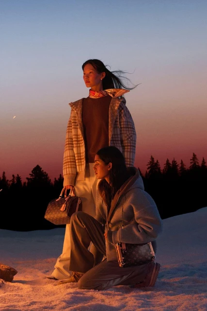 Two women in outerwear pose outdoors at twilight in a snowy landscape, carrying Louis Vuitton handbags with evergreen trees in the distance.