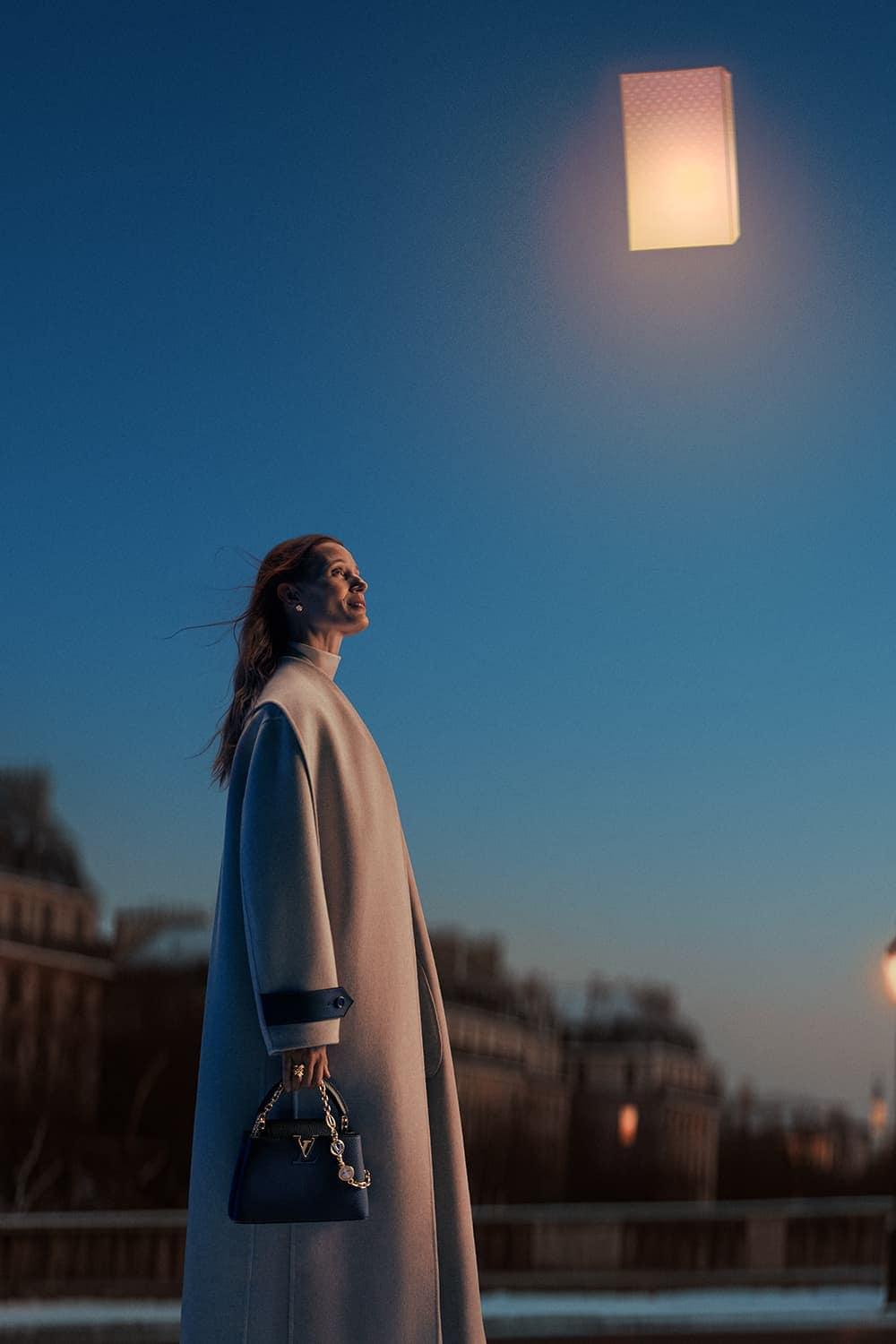Elegant woman in a coat carrying a handbag, with a lantern floating overhead in an outdoor, urban setting at dusk.