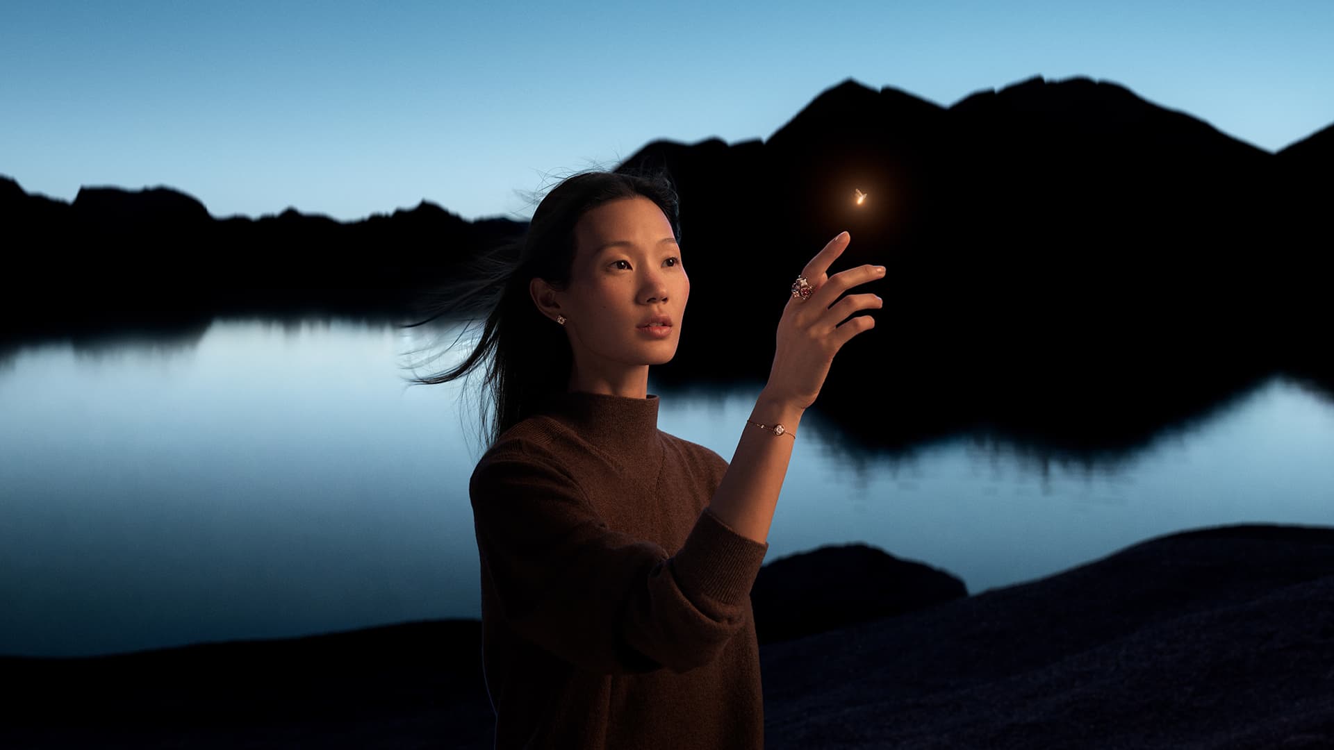 Portrait of a graceful young Asian woman outdoors in a brown turtleneck, gazing at a floating light in her open hand with lake backdrop.