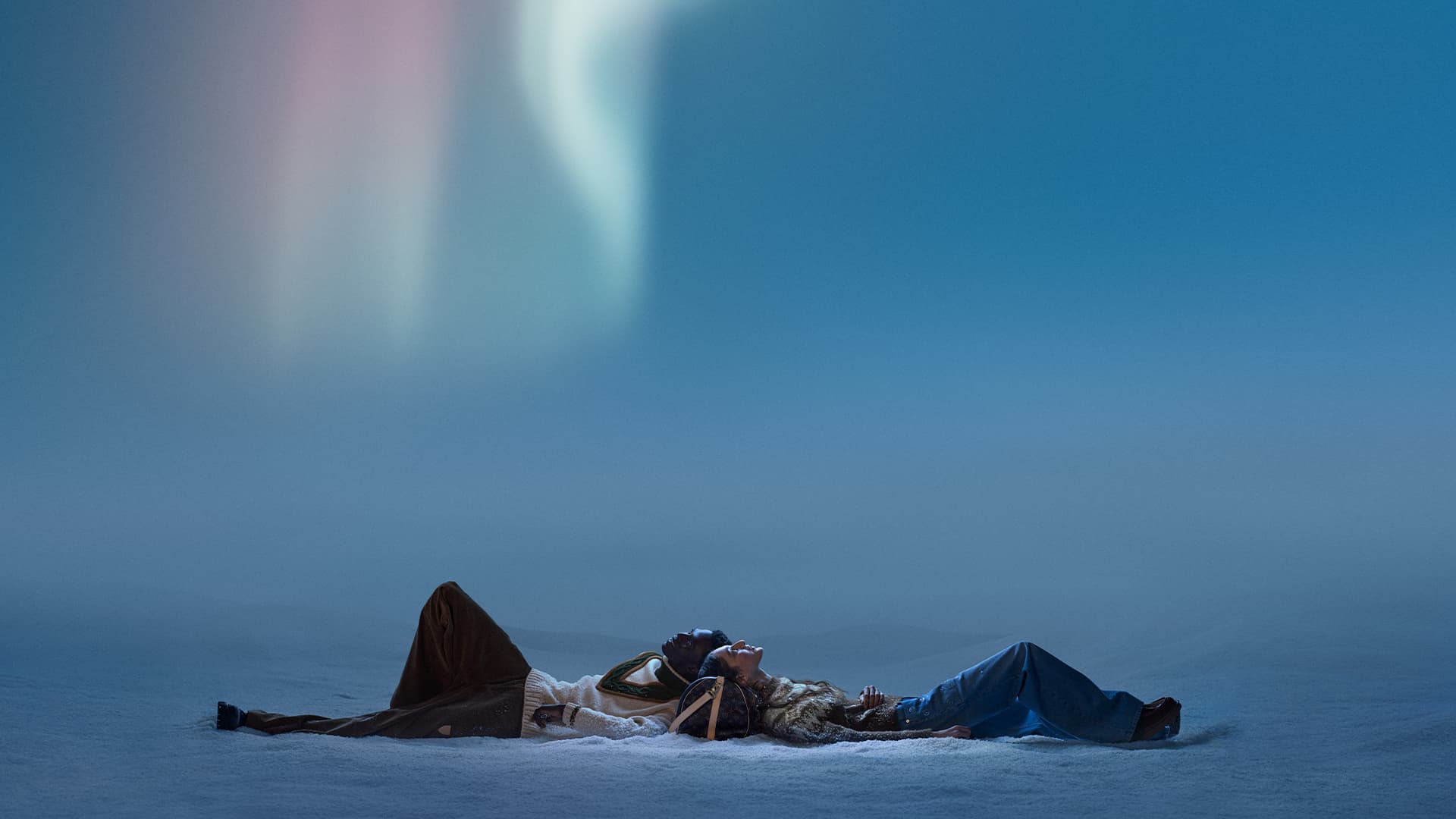 Two adults lie on snow, gazing up at the Northern Lights, wearing warm clothes and carrying a Louis Vuitton bag, against a blue sky.