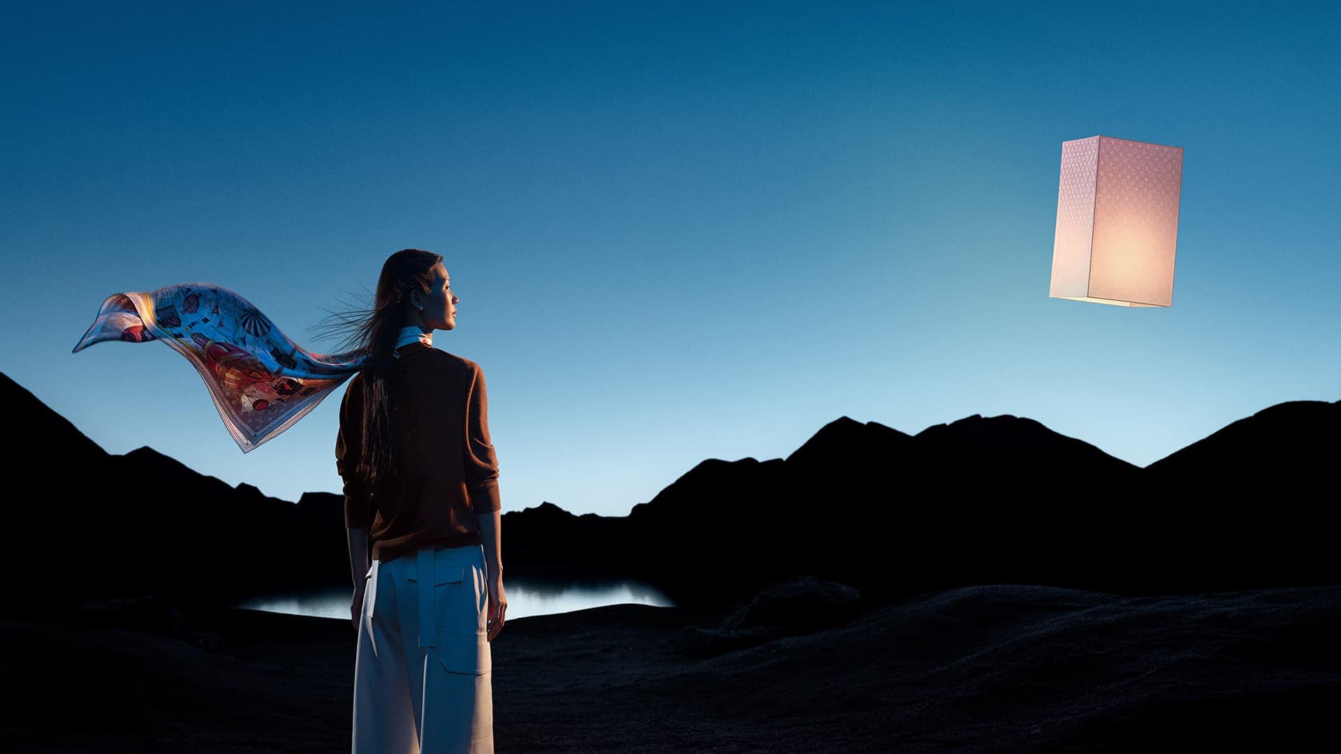 Woman gazes at illuminated lamp in scenic outdoor landscape at twilight, wearing scarf and brown sweater, mountains in the background.