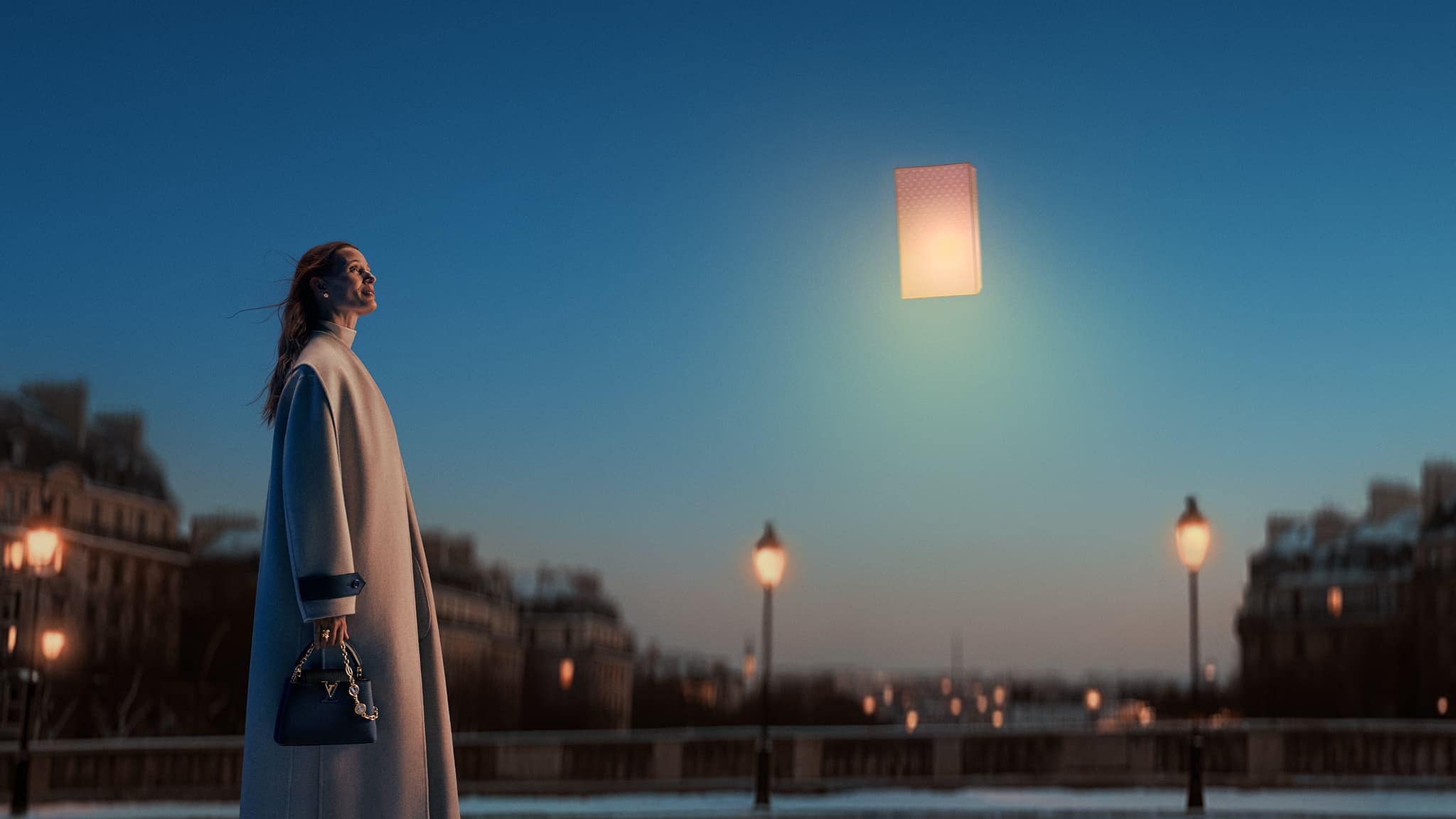 Elegant woman in coat stands by river at dusk, holding Louis Vuitton handbag with glowing box floating above. Paris buildings blurred.