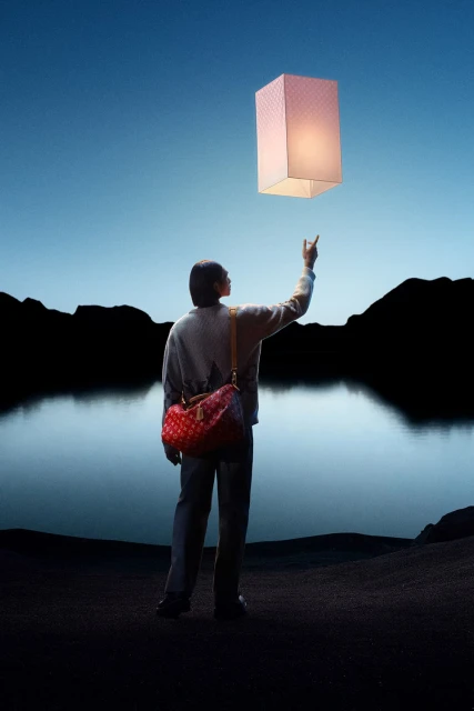 Adult reaches for glowing lantern in a serene nighttime landscape with a dark lake and mountain silhouettes under a clear twilight sky.