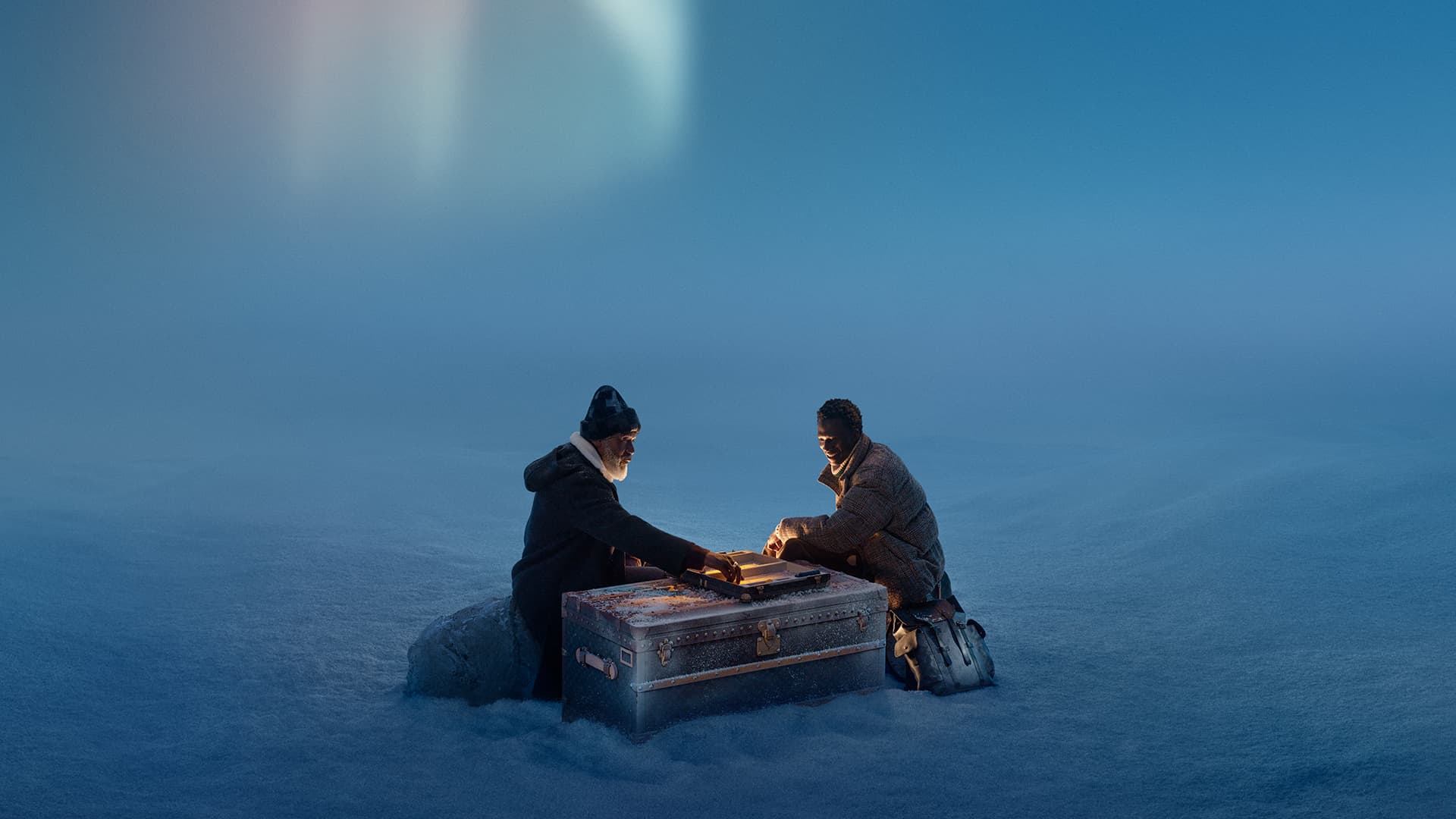 Two men sit in a snowy landscape beside a trunk with glowing edges. One man is older with a beard, and the other is younger.