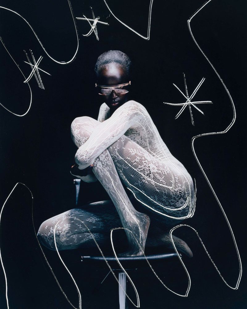 Model poses on a chair in lace bodysuit and futuristic sunglasses against a dark backdrop with abstract white light painting.