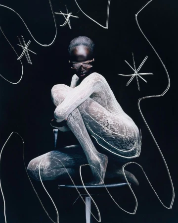 Model poses on a chair in lace bodysuit and futuristic sunglasses against a dark backdrop with abstract white light painting.