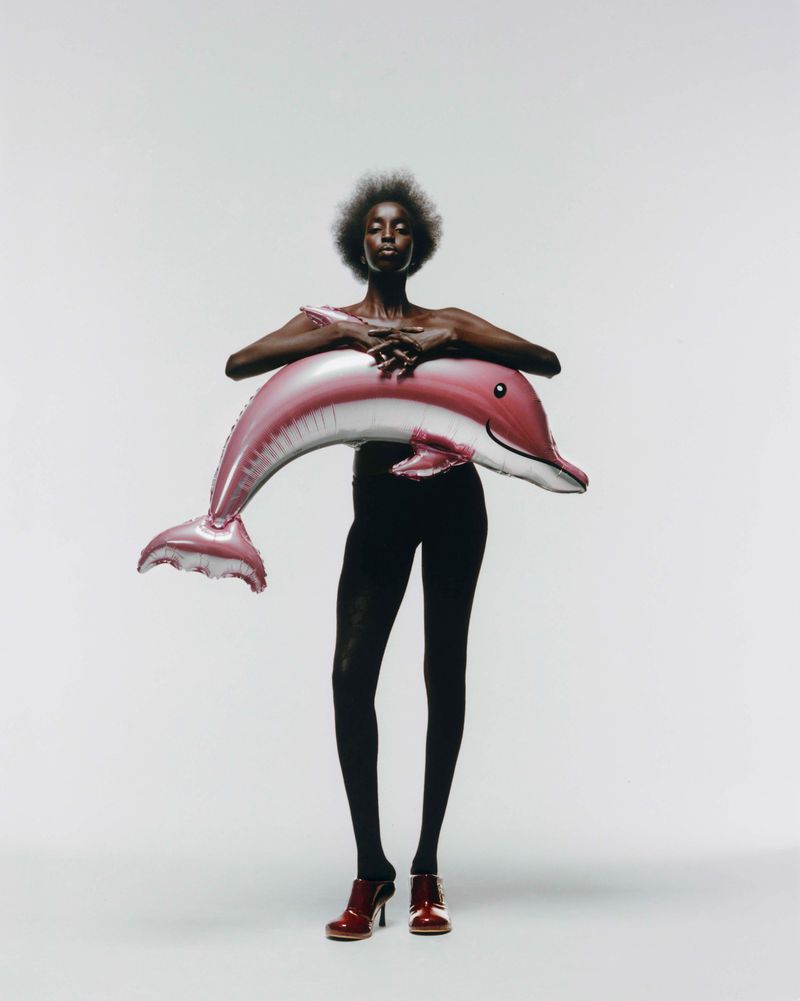 Full shot of an adult female with dark skin holding a pink dolphin balloon in a photo studio, wearing dark leggings and dark red mules.