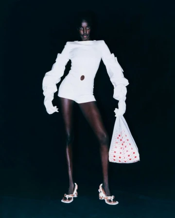 A statuesque Black fashion model strikes a pose against a black backdrop, showcasing a stylish white romper, sandals, and a polka-dot bag.