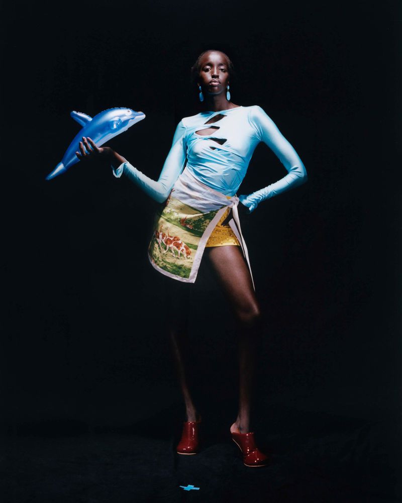 A statuesque Black model stands in a studio, light blue top with cutouts, decorative wrap skirt, red shoes, holding a blue toy dolphin.
