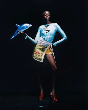 A statuesque Black model stands in a studio, light blue top with cutouts, decorative wrap skirt, red shoes, holding a blue toy dolphin.