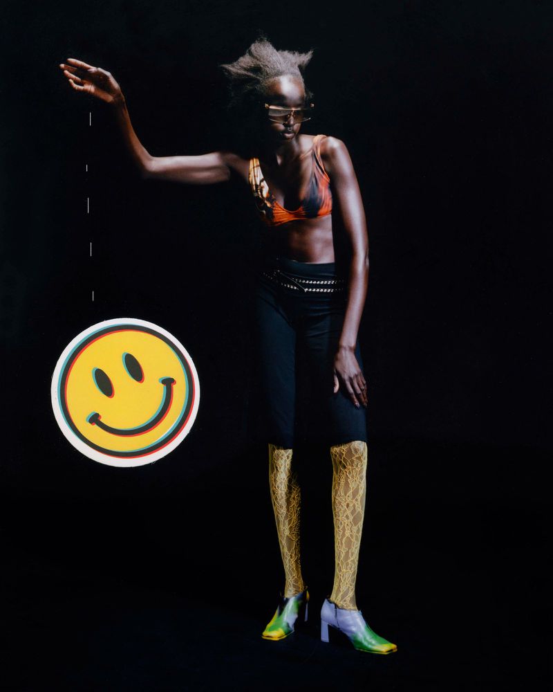 Fashion shot featuring a slender dark-skinned adult woman in a bra top, black pants, textured stockings, and heels, posing with a smiley-face prop.