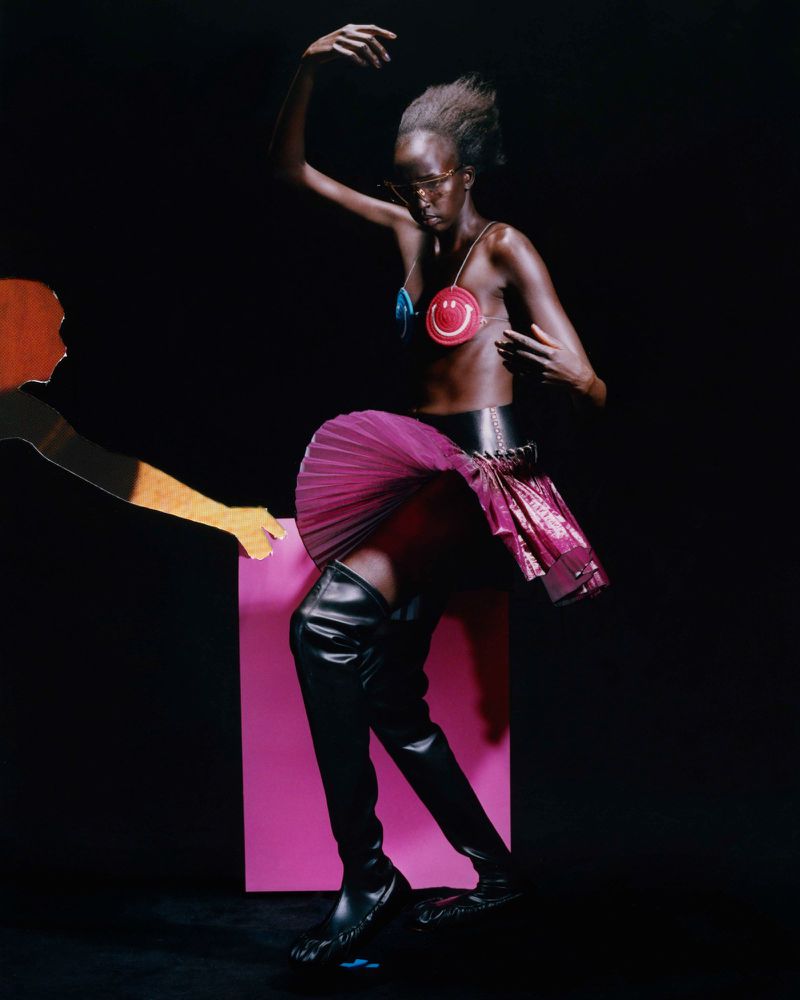 A striking editorial fashion shot of a slender dark-skinned woman in a unique outfit, balancing on a pink cube in a dark studio setting.