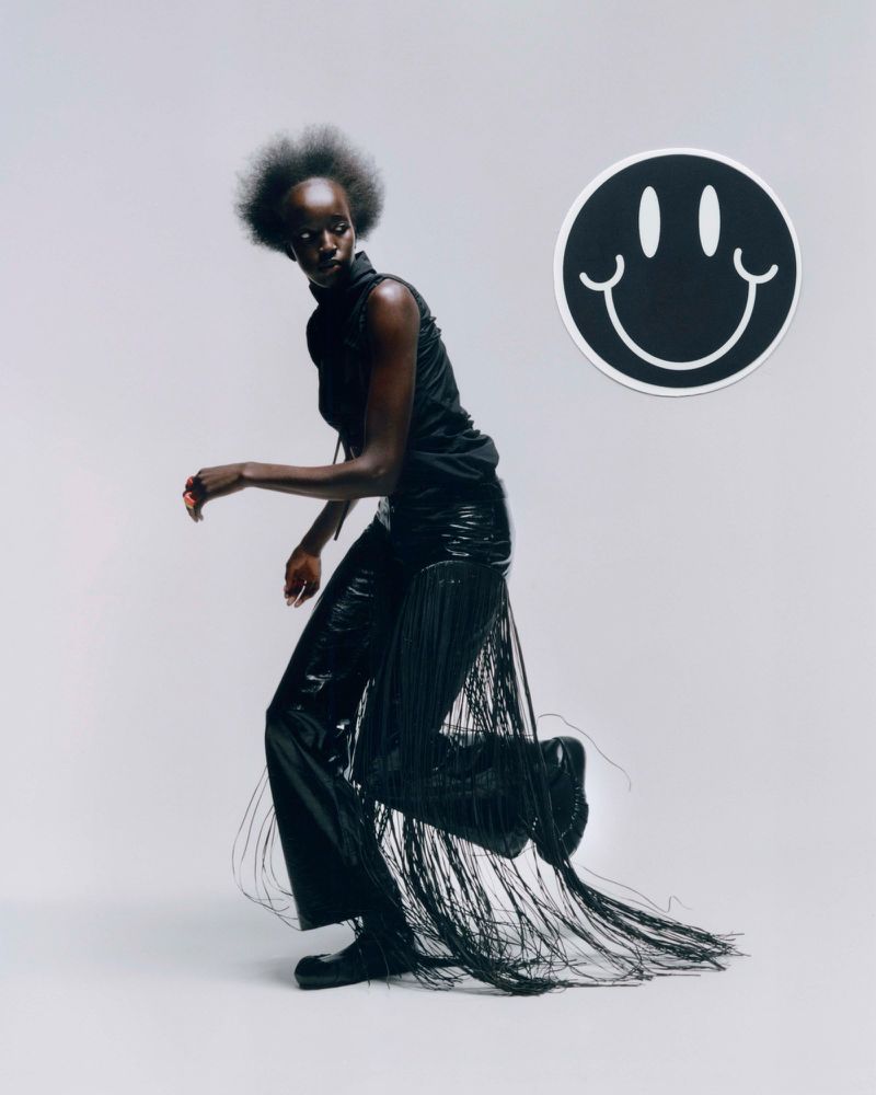 Fashion shot of a Black woman wearing black fringe pants and top in a light grey studio setting next to a smiley face decoration.
