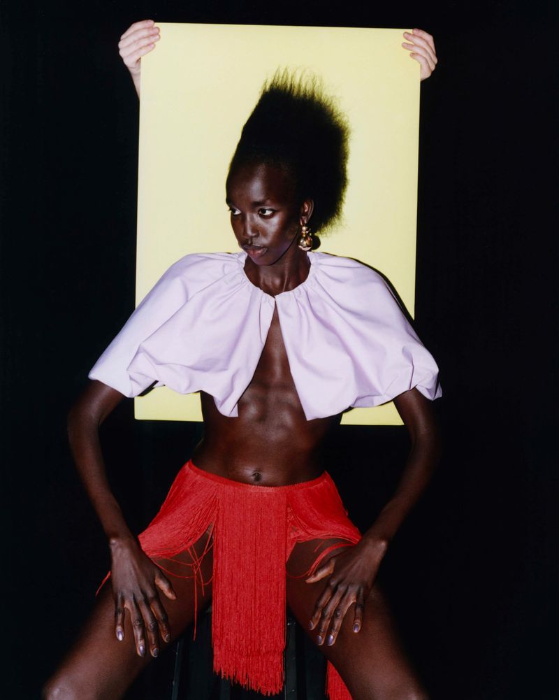Stunning studio portrait of an African female model with a dramatic fringe skirt, an open-front blouse, and hands holding a yellow screen.