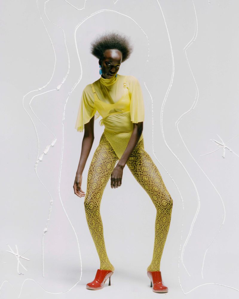 A Black female fashion model poses in a yellow bodysuit, printed leggings, and red heeled shoes against a bright white studio backdrop.