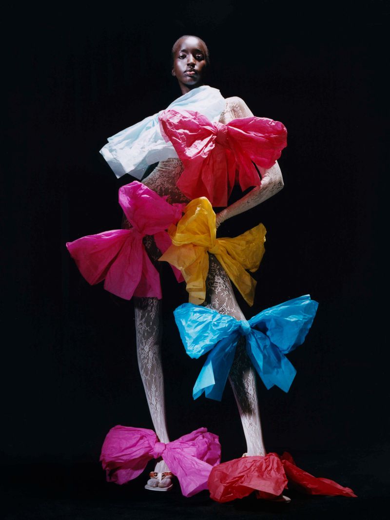 Fashion shot of a Black woman in a sculptural lace and colorful tissue-paper bow dress against a black background in a studio.