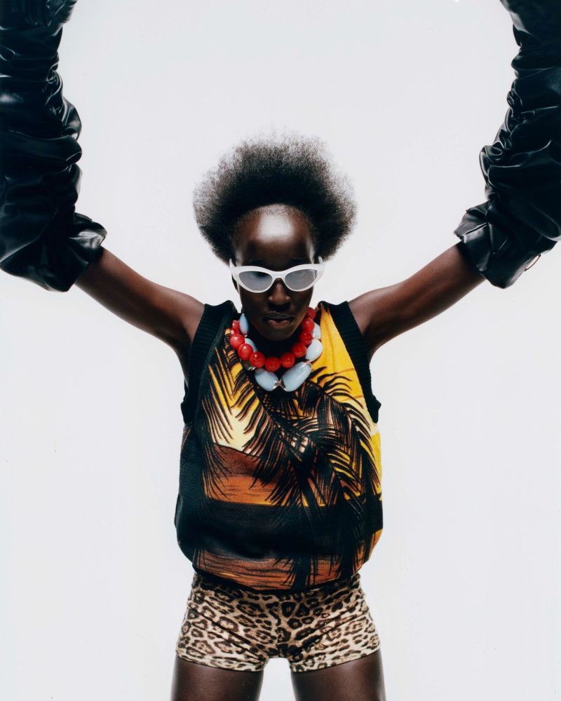 A striking fashion portrait of a young adult Black woman in sunglasses, statement jewelry, and printed top, against a neutral background.