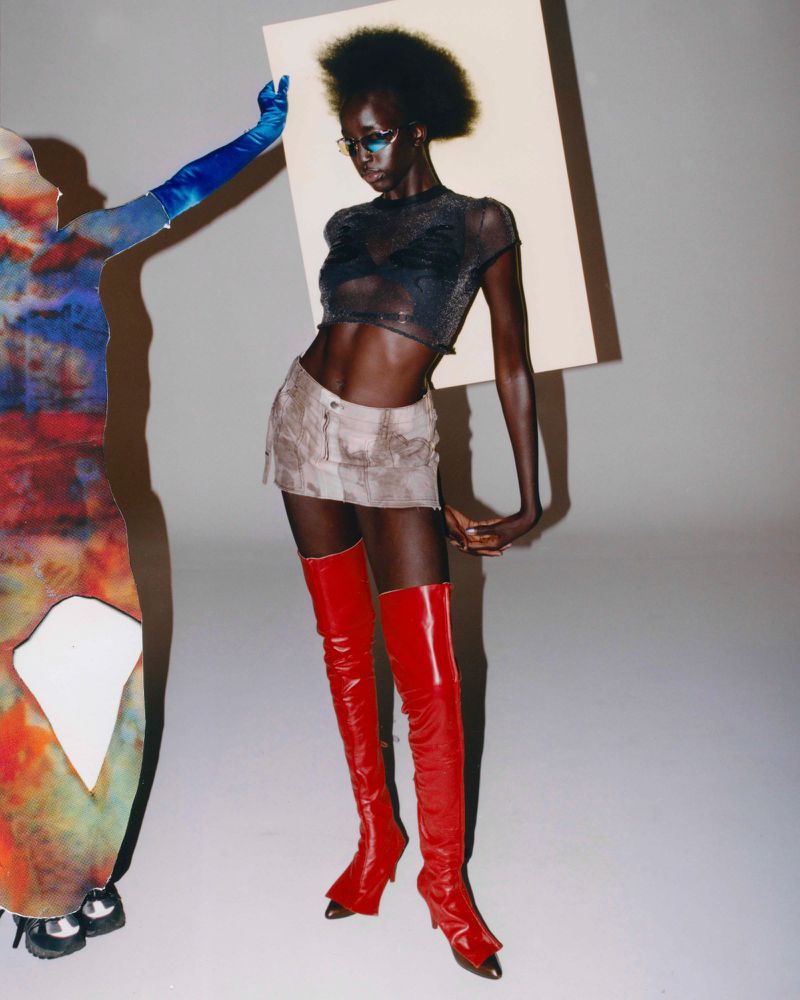 A fashion shot of a Black female model in a see-through top, miniskirt, and red boots posing next to a figure holding a white board.