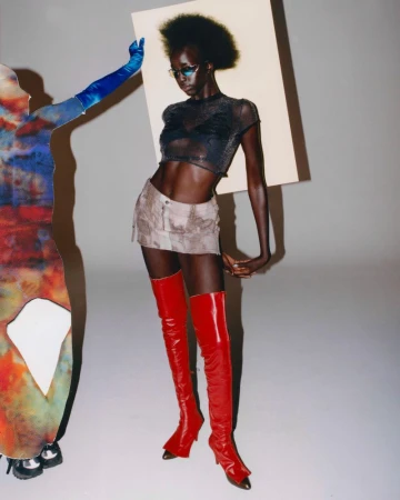 A fashion shot of a Black female model in a see-through top, miniskirt, and red boots posing next to a figure holding a white board.