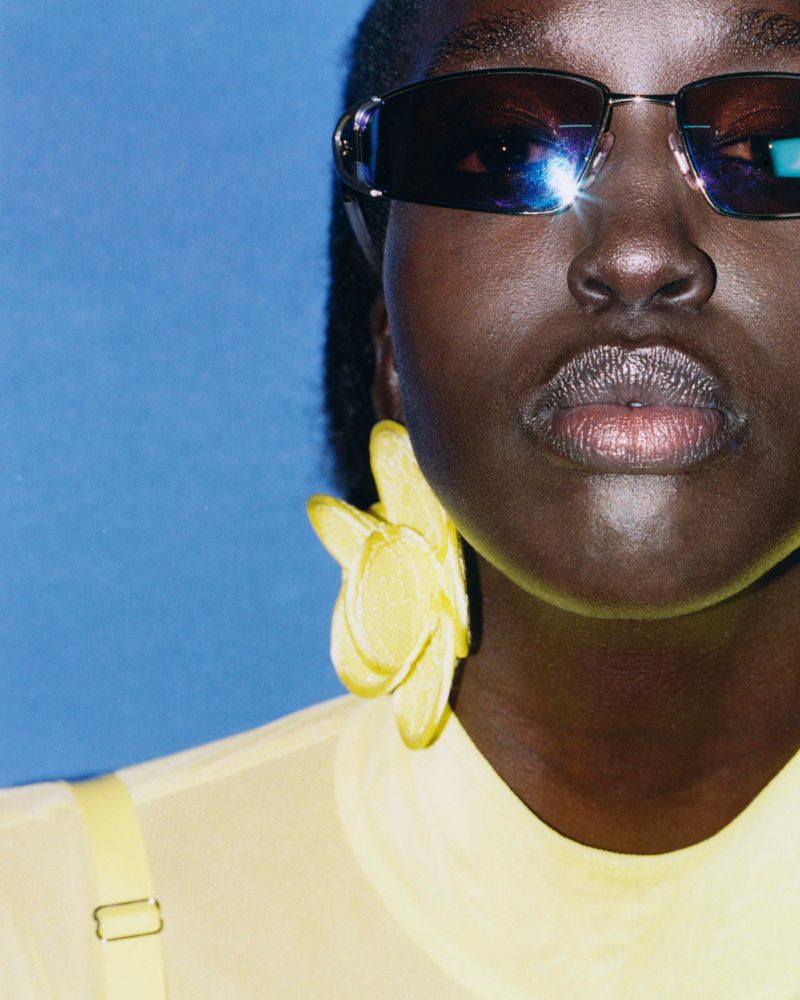 Close-up of an African woman with dark sunglasses and a large yellow flower earring against a solid blue background in studio lighting.
