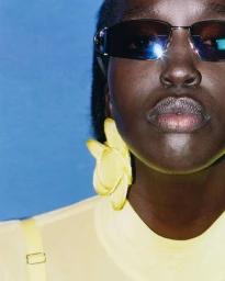 Close-up portrait of model Nyaueth Riam wearing dark sunglasses, yellow top, and flower earring against blue background