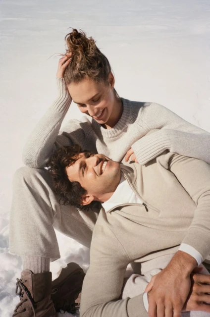 Couple poses smiling in outdoor winter scene with snow. Woman rests her head on man's chest, both wear neutral sweaters and pants.