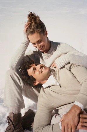 Couple poses smiling in outdoor winter scene with snow. Woman rests her head on man's chest, both wear neutral sweaters and pants.