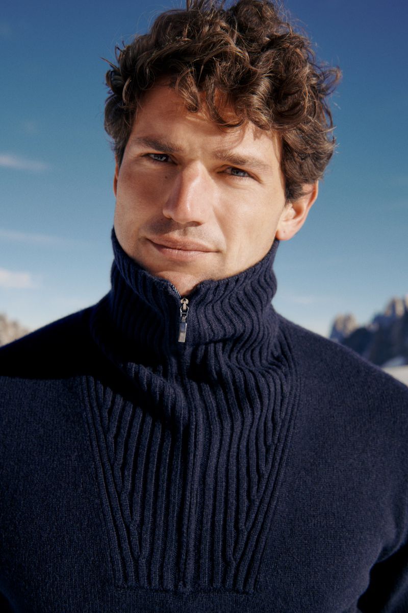 Man with curly brown hair wearing a navy blue ribbed quarter-zip sweater outdoors against a bright blue sky and mountain backdrop.