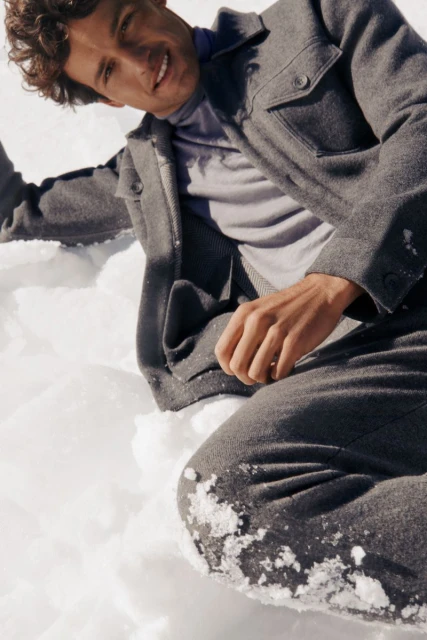 Smiling man in a gray jacket and pants sits in the snow, angled medium shot, outdoors, neutral tone, fashion or lifestyle content.