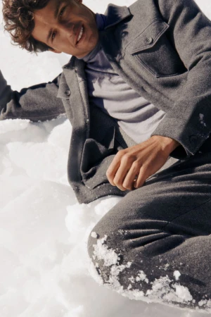 Smiling man in a gray jacket and pants sits in the snow, angled medium shot, outdoors, neutral tone, fashion or lifestyle content.