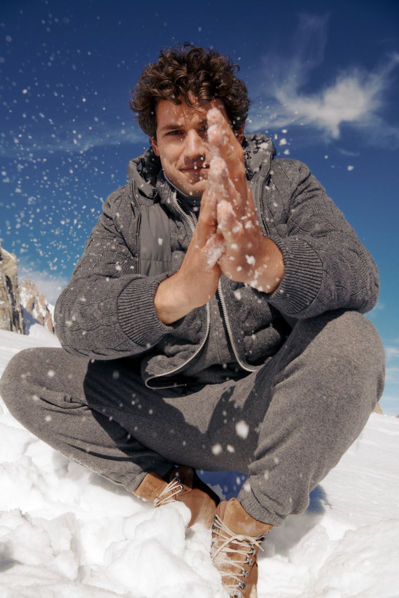 Attractive male model in gray knitwear sits cross-legged in snow against a blue sky and mountain backdrop, playfully throwing snow.
