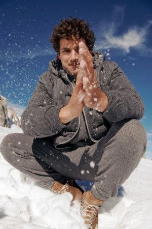 Attractive male model in gray knitwear sits cross-legged in snow against a blue sky and mountain backdrop, playfully throwing snow.
