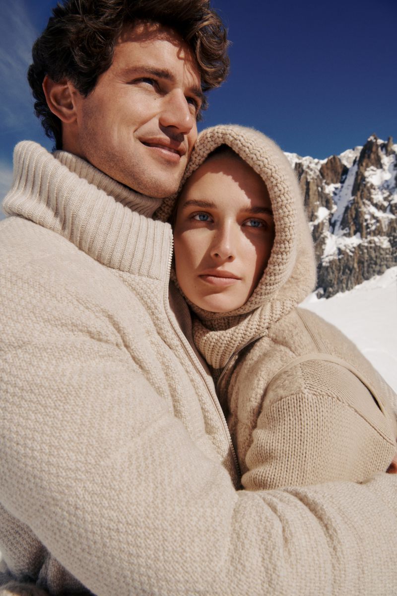 Close-up of a stylish couple in matching sweaters, embracing against a backdrop of snowy mountains and a clear blue sky.