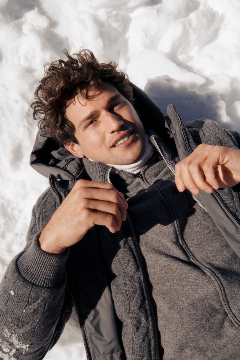 Man in gray hooded jacket smiles as he lies on his back in snow, directly facing the camera with a high-angle view in natural light.