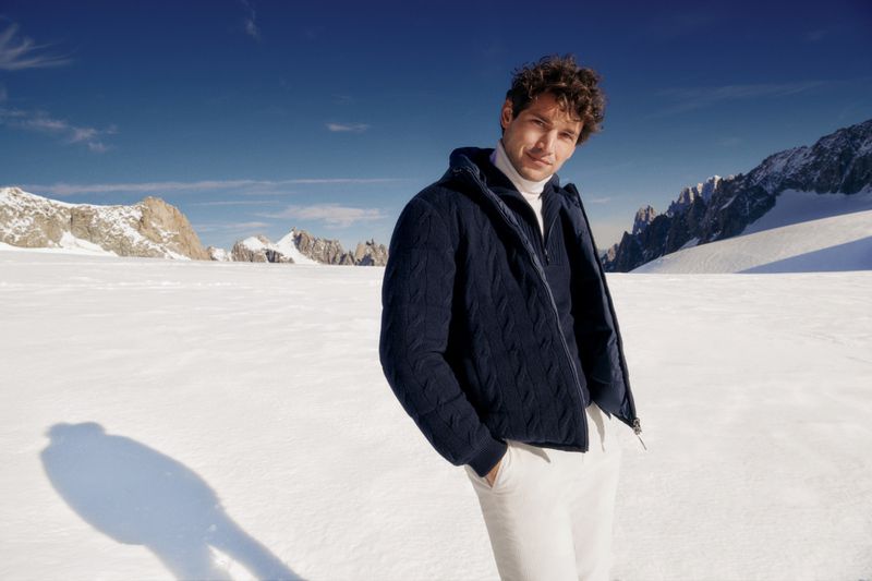Man in a blue puffer jacket and white pants stands against a backdrop of snow and mountains under a bright blue sky.